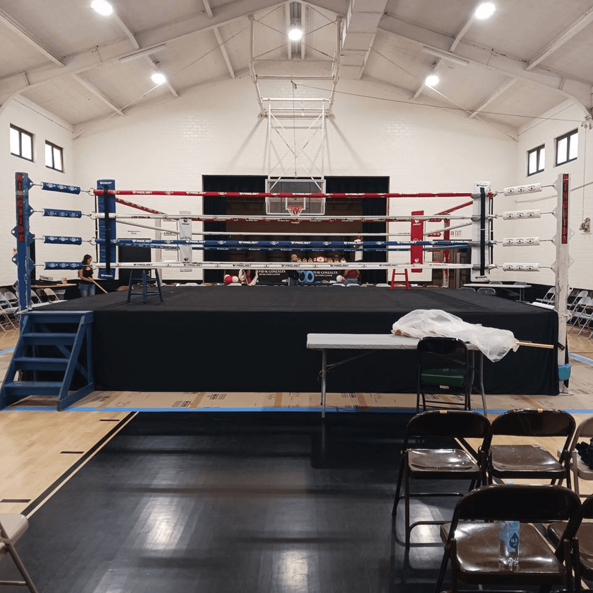 PROLAST 8 x 8 ft boxing ring, 1 ft platform: black-skirted ring with blue steps set up in a gymnasium with folding chairs
