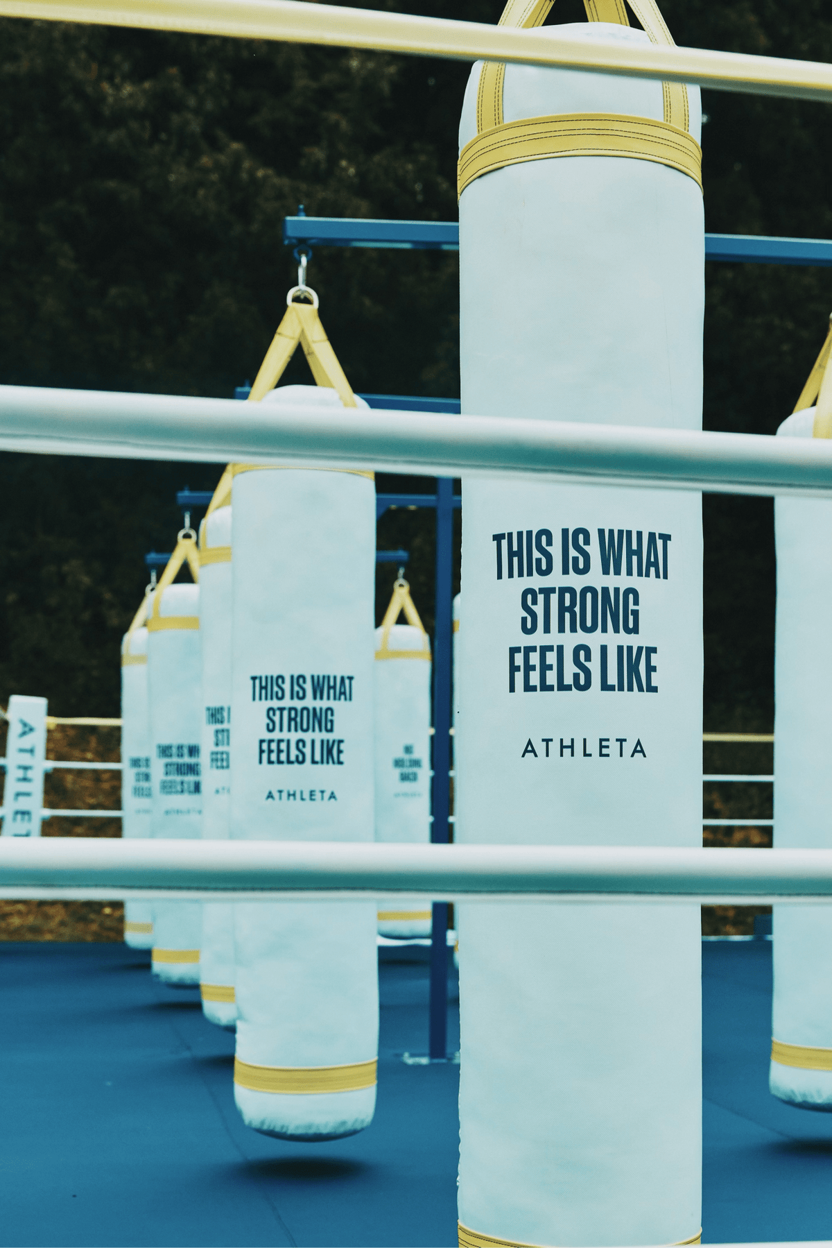PROLAST 10 x 10 ft boxing ring, 1 ft platform: white sponsor-branded heavy bags hanging behind the ring ropes outdoors