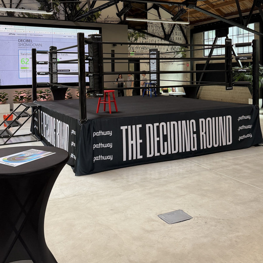 PROLAST boxing ring with black ropes, a red corner stool and ring steps, its printed skirt seen from a corner in an event hall
