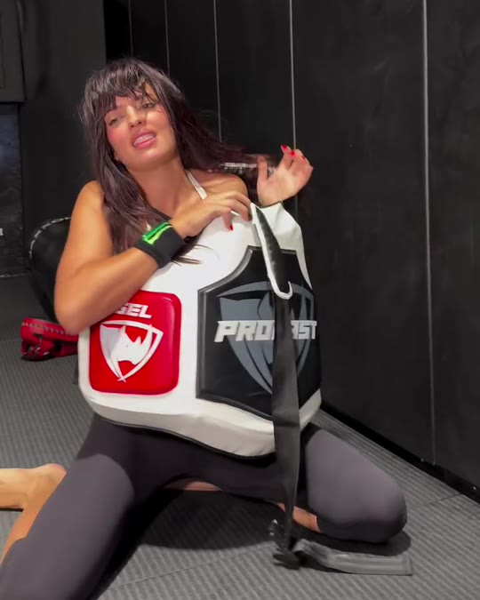 Nina Drama kneeling in a gym right after the kick, holding the PROLAST body protector, its logo in view